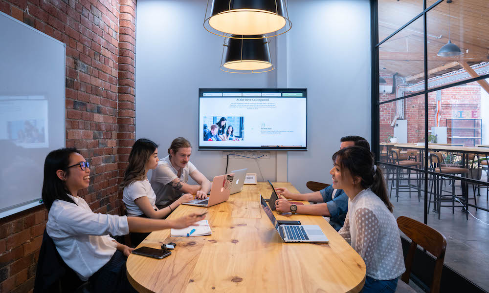 A group of 5 trendy professionals gathered around a large wooden table, within a good sized meeting room with exposed brick wall and warm lighting. They are working off of laptops and also have a presentation on display on a TV at the end of the Table.